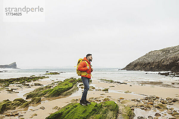 Mann mit Rucksack steht auf einem Felsen am Strand