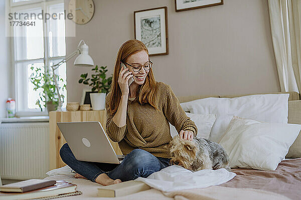 Lächelnde Frau telefoniert mit ihrem Smartphone und streichelt ihren Hund im heimischen Schlafzimmer