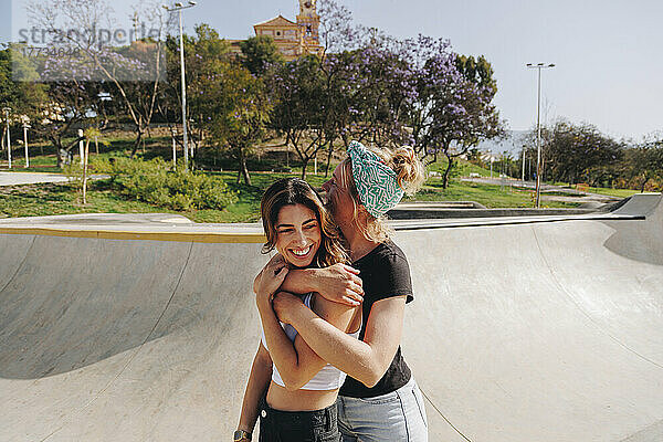 Happy woman embracing friend from behind at skateboard park on sunny day