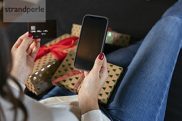 Frau mit Kreditkarte beim Online-Shopping per Smartphone sitzt neben Weihnachtsgeschenk