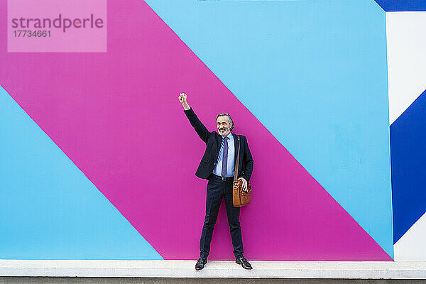 Happy businessman with arm raised standing in front of pink and blue wall