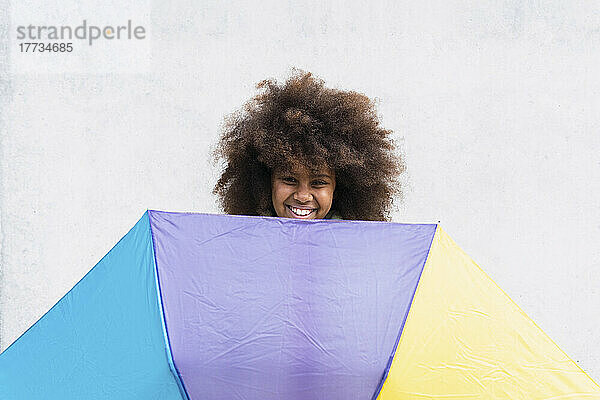 Lächelndes Mädchen mit buntem Regenschirm vor weißer Wand