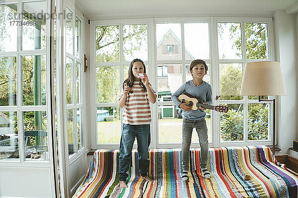 Geschwister spielen Musikinstrument und stehen zu Hause auf dem Sofa