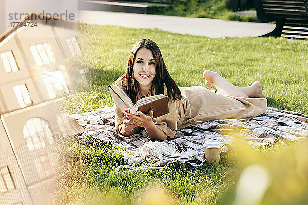 Schöne lächelnde Frau mit Buch  die an einem sonnigen Tag im Park liegt
