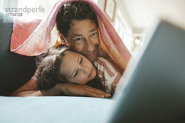 Smiling mature man with daughter lying under blanket at home