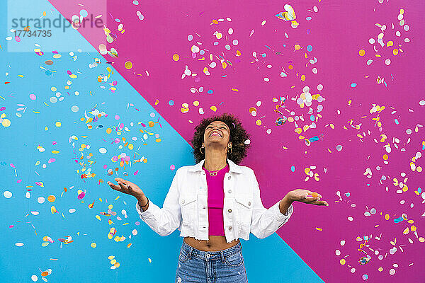 Glückliche Frau mit Afro-Frisur steht inmitten von Konfetti vor einer rosa und blauen Wand