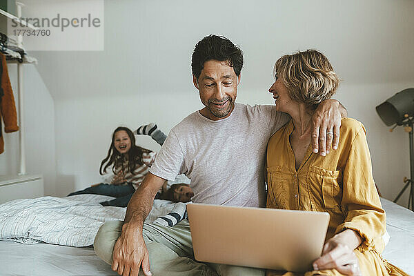 Mann mit Laptop sitzt neben Frau und schaut Tochter und Sohn zu  die zu Hause im Bett spielen