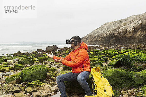 Mann mit Virtual-Reality-Simulator gestikuliert auf einem Felsen sitzend