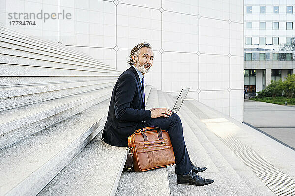 Lächelnder Geschäftsmann mit Laptop sitzt auf Stufen