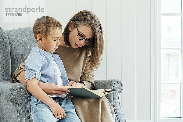 Mutter und Sohn lesen Buch und sitzen zu Hause im Sessel