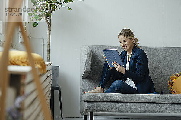 Happy businesswoman looking at tablet PC sitting on sofa in office