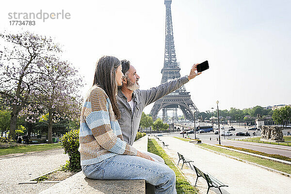 Reifer Mann macht Selfie mit Frau per Smartphone  die an der Wand vor dem Eiffelturm sitzt  Paris  Frankreich