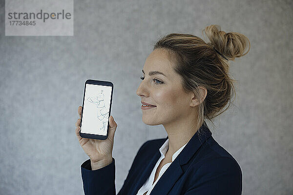 Lächelnde Geschäftsfrau zeigt Smartphone-Bildschirm vor grauer Wand