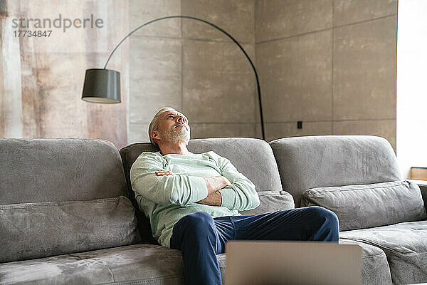Thoughtful mature man with arms crossed sitting on sofa at home