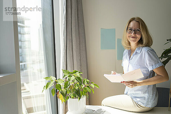 Lächelnder Freiberufler mit Brille sitzt mit Tagebuch auf dem Schreibtisch im Heimbüro