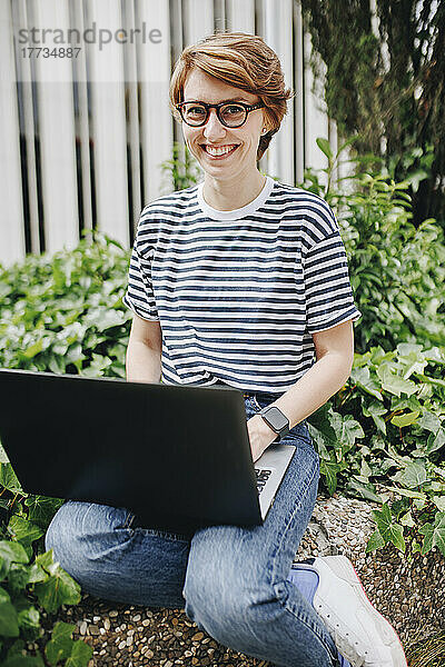Glückliche Frau mit Laptop sitzt an der Wand neben Pflanzen