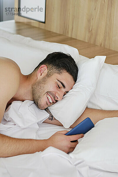 Glücklicher Mann mit Mobiltelefon  der zu Hause im Bett aufwacht