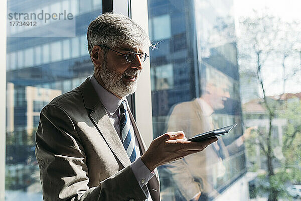 Glücklicher Geschäftsmann mit Smartphone am Fenster im Büro
