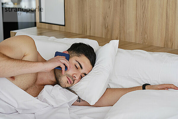 Young man talking through mobile phone lying in bed at home