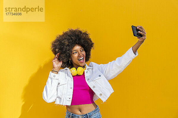 Glückliche junge Frau macht ein Selfie mit dem Handy vor einer gelben Wand