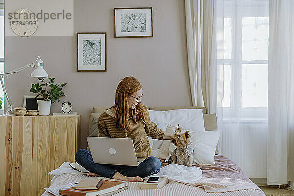 Lächelnde Frau sitzt mit Laptop und streichelt ihren Hund zu Hause im Bett