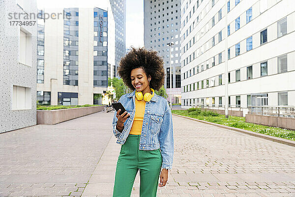 Glückliche Afro-Frau mit kabellosen Kopfhörern und Mobiltelefon  die auf dem Fußweg steht