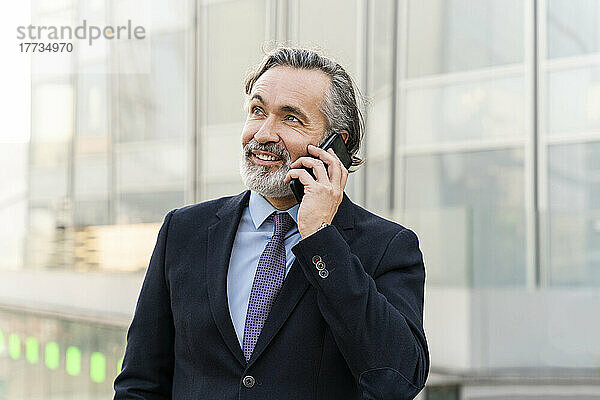 Lächelnder reifer Geschäftsmann mit grauen Haaren  der auf dem Smartphone spricht
