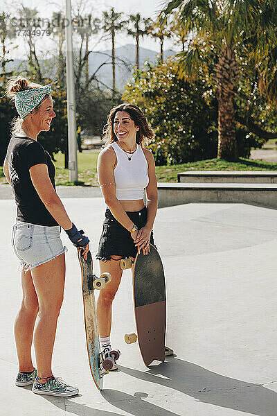 Lächelnde Freunde halten Skateboards und unterhalten sich an einem sonnigen Tag auf der Sportrampe