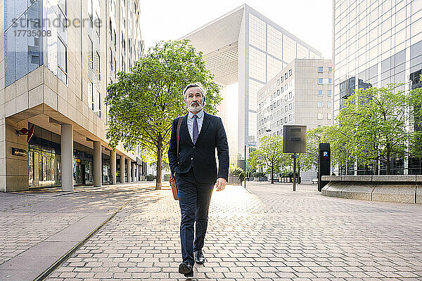 Reifer Geschäftsmann mit Tasche spaziert an sonnigem Tag im Büropark