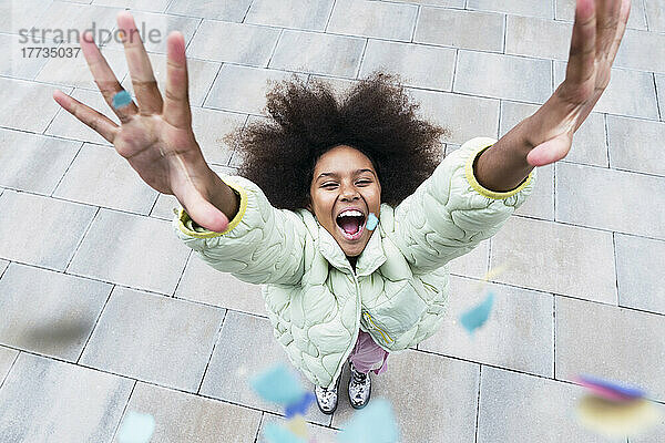 Cheerful girl with arms raised standing amidst confetti falling on footpath