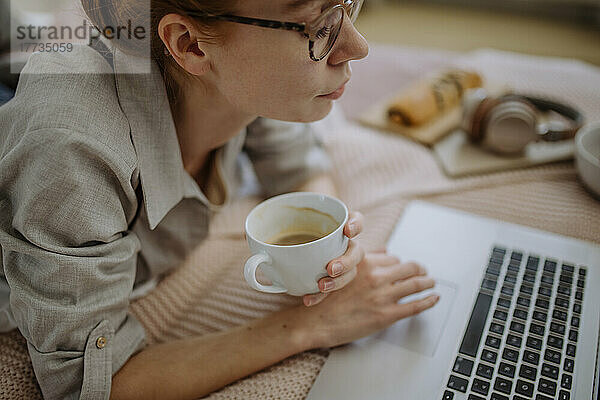 Frau mit Brille und Kaffeetasse hält zu Hause E-Learning über Laptop