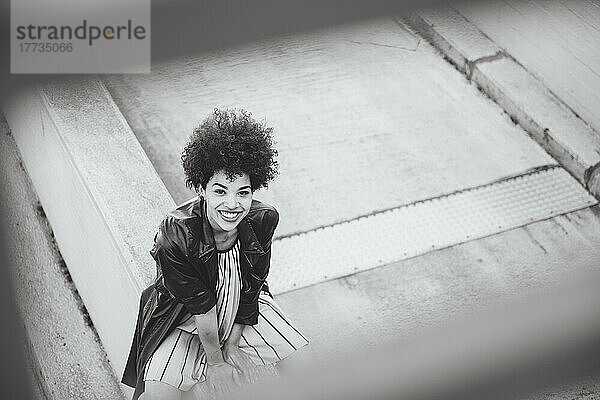 Fröhliche junge Frau mit Afro-Frisur sitzt an der Wand
