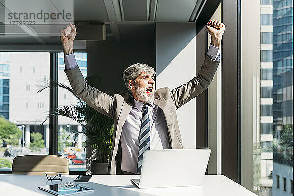 Glücklicher Geschäftsmann feiert Erfolg am Schreibtisch im Büro
