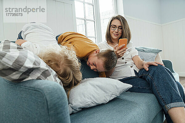 Glückliche Frau benutzt Smartphone von Söhnen  die zu Hause auf dem Sofa spielen