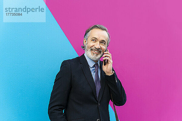 Glücklicher Geschäftsmann  der mit seinem Smartphone spricht und vor einer rosa und blauen Wand steht