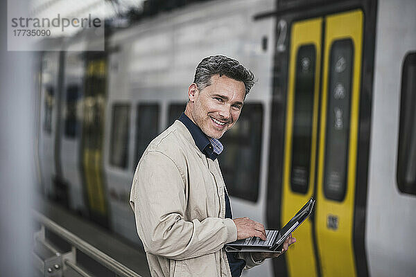 Glücklicher Geschäftsmann mit Tablet-PC  der vor dem Zug am Bahnhof steht