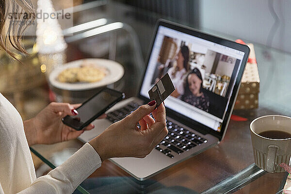 Frau mit Kreditkarte und Smartphone führt zu Hause Videoanrufe mit Freunden über Laptop