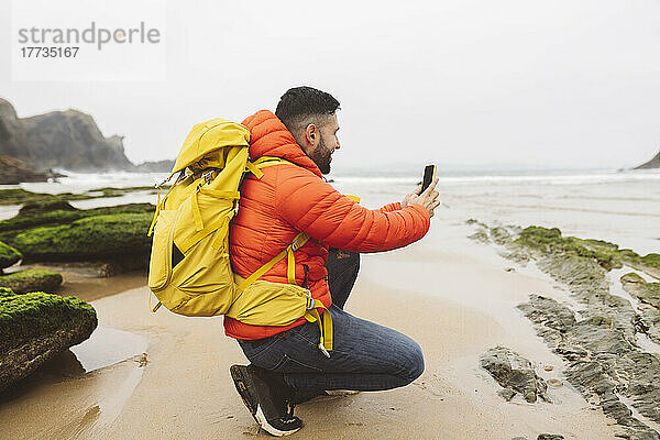 Mann mit Rucksack fotografiert am Strand mit Smartphone