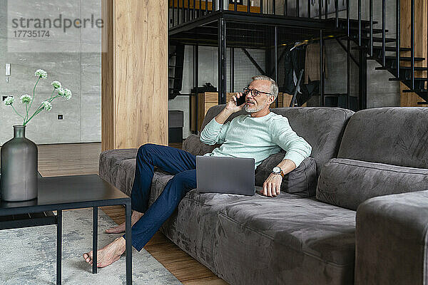 Geschäftsmann spricht mit Smartphone und sitzt mit Laptop auf dem heimischen Sofa