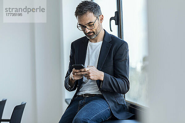 Geschäftsmann  der Textnachrichten über sein Smartphone sendet  sitzt auf der Fensterbank im Büro