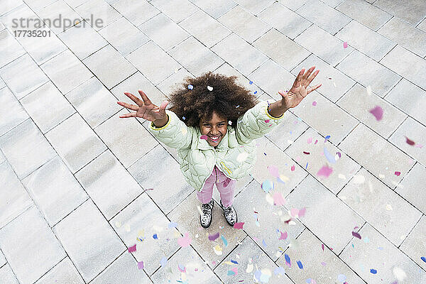 Cheerful Afro girl with arms raised standing amidst confetti falling on footpath
