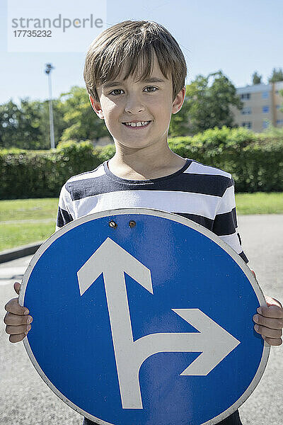 Lächelnder Junge hält an einem sonnigen Tag ein Verkehrsschild mit Pfeilsymbol