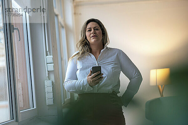 Selbstbewusste Geschäftsfrau mit Hand an der Hüfte und Smartphone im Büro