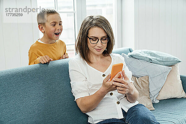 Fröhlicher Junge schaut Mutter zu  die zu Hause auf dem Sofa ihr Smartphone benutzt