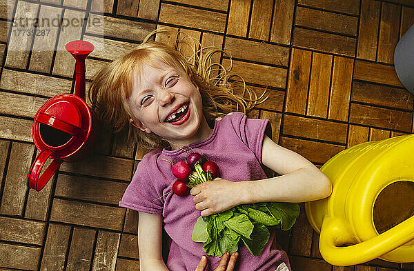 Cheerful girl with radish lying by watering can on balcony floor