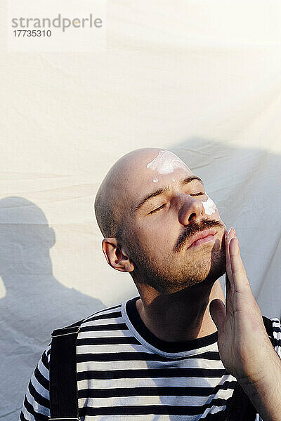 Glatzköpfiger Mann mit geschlossenen Augen  der vor weißem Hintergrund Feuchtigkeitscreme aufträgt