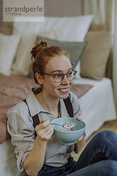 Glückliche Frau mit Brille sitzt zu Hause mit einer Schüssel Essen vor dem Bett