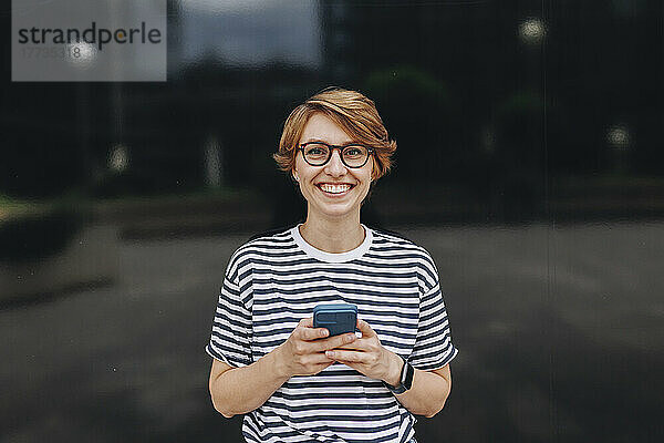 Lächelnde Frau mit Brille und Smartphone vor schwarzer Wand