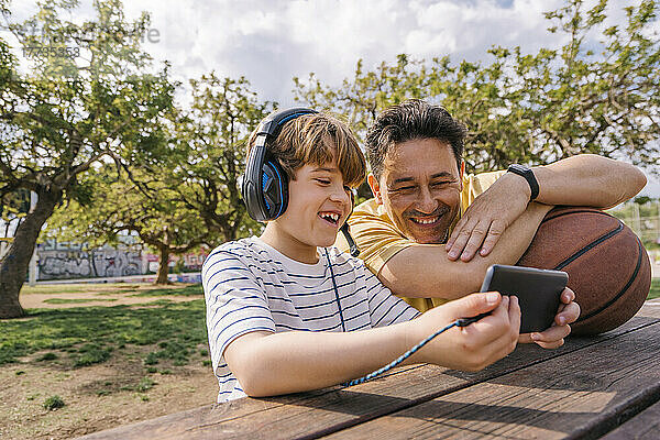 Lächelnder Junge hört Musik über Kopfhörer und teilt sein Mobiltelefon mit seinem Vater  der am Tisch sitzt