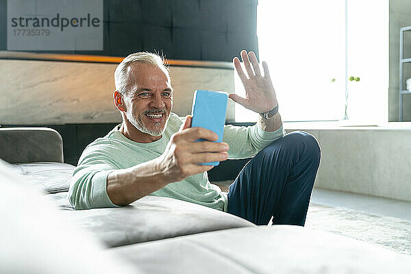 Lächelnder Mann winkt per Videoanruf seinem Smartphone im Wohnzimmer zu